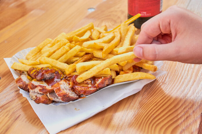 Hand greift zu Pommes mit Currywurst in einer Pappschale mit einer Cola daneben.