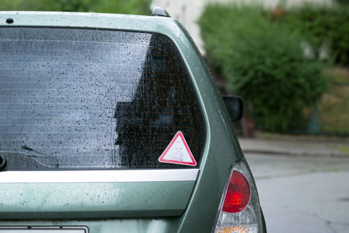 Auf der Rückscheibe eines Autos, ein gewöhnlicher PKW, ist ein Aufkleber zu sehen. Es ist ein Warndreieck rot und weiß und soll Autofahrer zur Vorsicht ermahnen und die Aufmerksamkeit des hinteren Fahrers fördern.