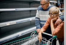 Eine ältere Frau und ein älterer Mann stehen vor einem leeren Regal in einem Supermarkt. Sie sind offenbar ein Paar. Der Mann legt seine Hand über die Schulter der Frau. Die beiden sehen bestürzt und besorgt aus. 