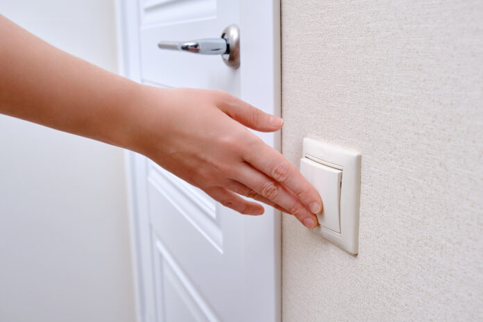 Hand reicht nach einem Lichtschalter neben einer weißen Tür mit einer silbernen Türklinke.