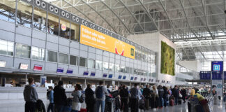 Ein Flughafen, an dem unzählig viele Passagiere und Reisende in einer Schlange warten. Sie haben Gepäck dabei und wollen fliegen. Doch offensichtlich gibt es Verspätungen oder Ausfälle. Die Halle des Flughafens ist groß und silbern. Man sieht Werbung, Counter und digitale Anzeigen zur Information.