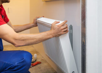 Ein Heizungsbauer installiert einen kleineren weißen Heizkörper vor einer weißen Wand. Zu sehen ist links der Anschnitt des Monteurs in blauer Hose und rotem Shirt.