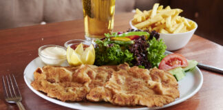 Ein Wiener Schnitzel befindet sich auf einem Teller auf einem Tisch in einem Restaurant. Neben dem Schnitzel steht eine Schale mit Zitronen und als Beilage gibt es einen gemischten Salat. Neben dem Teller steht eine Portion Pommes.