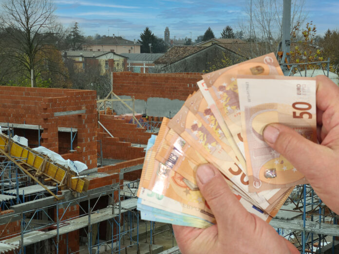 Eine Person hält mehrere Geldscheine in der Hand, im Hintergrund befindet sich ein Haus mitten im Bau