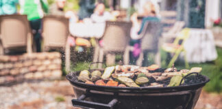 Ein Kugelgrill mit Grillgemüse auf dem Rost. Im Hintergrund findet eine Gartenparty statt.