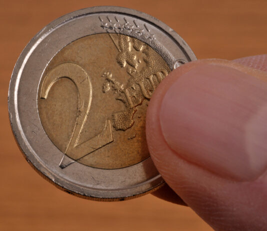 Eine 2-Euro-Münze wird in der Hand gehalten. Sie ist außen silber und innen Gold. Die Person hält sie zwischen Daumen und Zeigefinger.