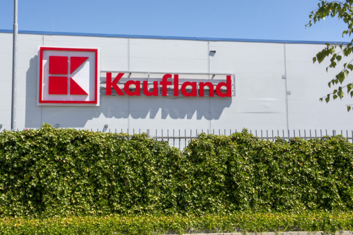 Ein große Kaufland-Filiale in einem großen Gebäude. Davor befindet sich eine hochgewachsene Hecke. Auf dem wei0en Gebäude befindet sich das rot-wei0e Kaufland-Logo sowie der rote Schriftzug "Kaufland"