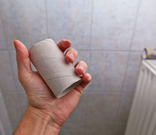 Eine Männerhand hält eine leere Toilettenpapier-Rolle in der Hand im Badezimmer