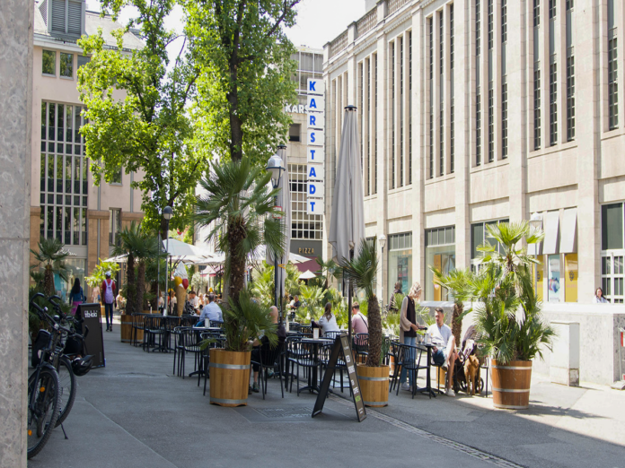 Seitenstraße in der Innenstadt von Karlsruhe. Blumentöpfe mit Pflanzen stehen auf dem Fußweg und verschönern die Passage. Im Hintergrund laufen Leute und schlendern durch die Innenstadt.