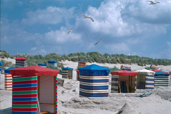 Viele bunte Strandkörbe stehen auf einem Sandstrand, vermutlich an der Nordsee. Es ist sonnig, Möwen fliegen am blauen Himmel. Im Hintergrund sind Dünen.