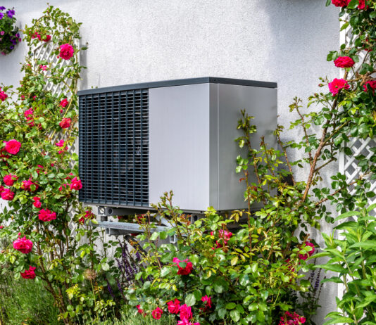 Wärmepumpe an der Hausfassade. Die Wärmepumpe steht direkt an der Wand umgeben von roten Rosen und erzeugt mittels Strom Wärme. Es handelt sich um ein fabrikneues Gerät ohne Gebrauchsspuren.