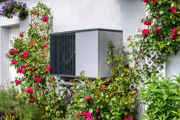 Wärmepumpe an der Hausfassade. Die Wärmepumpe steht direkt an der Wand umgeben von roten Rosen und erzeugt mittels Strom Wärme. Es handelt sich um ein fabrikneues Gerät ohne Gebrauchsspuren.