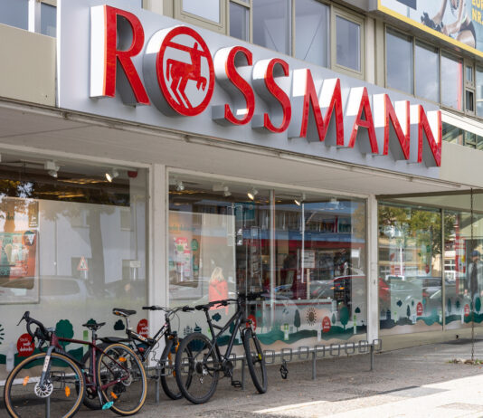 Der Eingangsbereich einer Rossmann-Filiale. Über der Eingangstür steht der Name der Drogerie und das Logo. Die Glastür öffnet sich automatisch und gibt den Blick auf das Sortiment und den Kassenbereich frei.