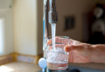 Eine Frau hält in einer Spüle ein Glas unter den fließenden Wasserhahn. Das Leitungswasser läuft heraus und sprudelt im Glas durch die Verwirbelungen. Im Hintergrund ist verschwommen die Küche.