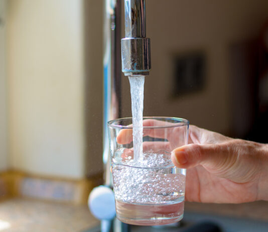 Eine Frau hält in einer Spüle ein Glas unter den fließenden Wasserhahn. Das Leitungswasser läuft heraus und sprudelt im Glas durch die Verwirbelungen. Im Hintergrund ist verschwommen die Küche.