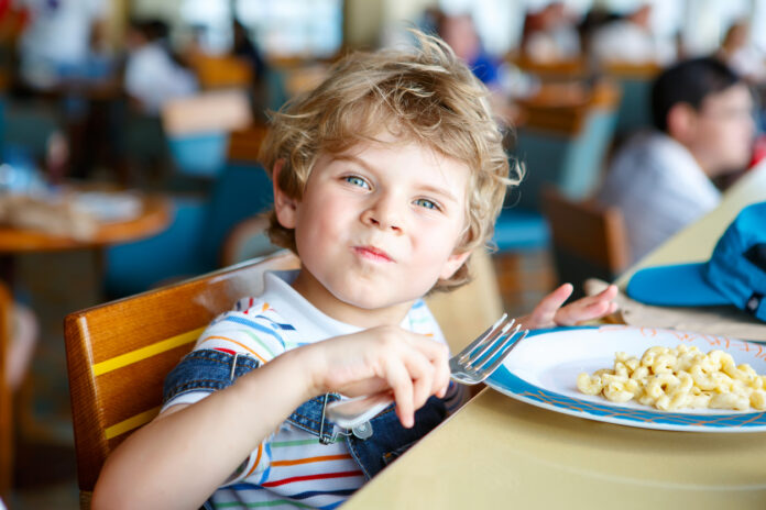 Ein süßer blonder Schuljunge isst sein Essen in der Schule und blickt mit der Gabel in der Hand in die Kamera.