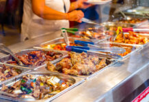Eine Person mit weißer Kleidung nimmt sich Essen aus einem Buffet. Es stehen viele verschiedene Gerichte auf einer Theke nebeneinander.