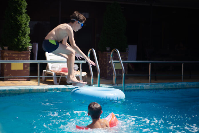 Ein Kind mit einer Badehose und einer Taucherbrille springt in ein Becken eines Schwimmbads. Daneben ist ein Junge mit Schwimmflügeln.