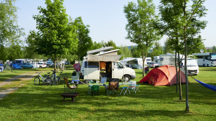 Ein schöner Campingwagen mit einem aufgebauten Dachzelt steht auf einer Wiese, auf einer großflächigen Fläche, die zu einem Campingplatz gehört. Daneben ist ein oranges Zelt zu sehen, so wie eine Camping-Frühstücksgarnitur.