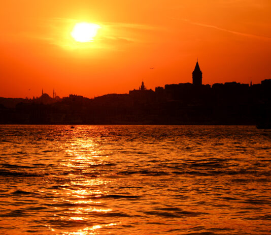 Die Sonne geht gerade über einer Stadt unter. Gegen den orangefarbenen Himmel zeichnet sich die Silhouette der Gebäude ab. Im Vordergrund färbt die Sonne das Wasser orange.