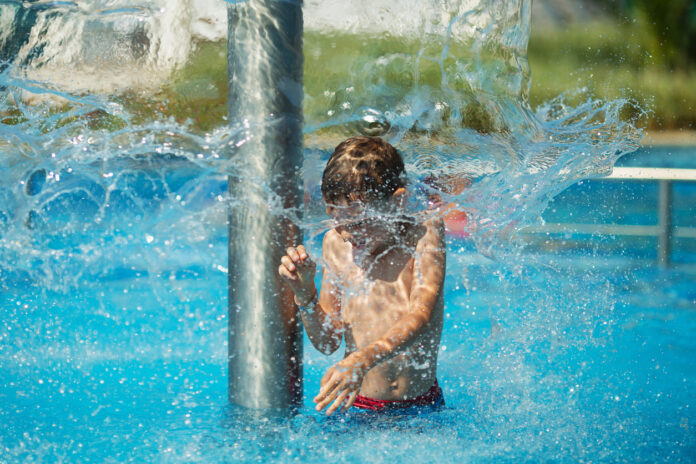 Ein Junge ist in einem Freibad und kühlt sich unter einer Fontäne ab. Er hat sichtlich Freude an dem Wasserspiel, während um ihn herum das Wasser in Bewegung ist. Der Pool strahlt in einem türkisen Blau.