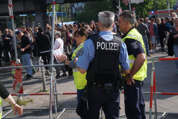 Polizisten und Mitarbeiter des Ordnungsamtes überwachen eine Demonstration oder eine Menschenansammlung auf der Straße. Sie tragen Warnwesten mit der Aufschrift "Polizei". Außerdem sind sie bewaffnet.