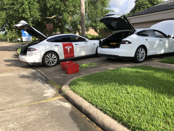 Zwei weiße E-Autos von Tesla stehen nebeneinander in einer Auffahrt. Ihre Kofferräume sind geöffnet. Am Straßenrand ist ein Vorgarten mit frischem grünen Gras.