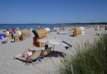 Viele Urlauber entspannen an einem weißen Sandstrand vor einem kristallklaren, blauem Meer vor blauem Himmel auf Decken und in diversen Strandkörben, die an der Küste verstreut sind und Schutz vor der Sonne bieten.