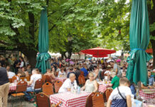 Viele gut gelaunte Menschen sitzen beim schönem Wetter draußen auf einer Terrasse oder in einem Biergarten eines Gasthauses und trinken und essen etwas in großer Gesellschaft und heiterer Stimmung.