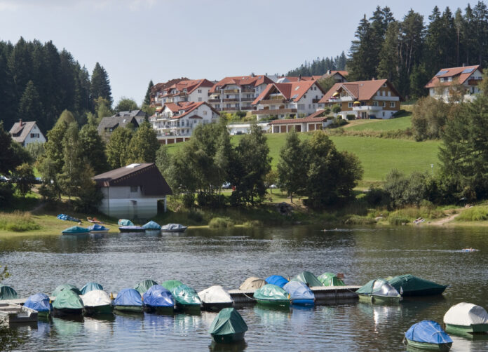 Ferienhäuser, Pensionen und Hotels in traditioneller Umgebung. Davor liegt ein idyllischer großer See oder Badesee mit mehreren Booten. Der Ort bzw. das Dorf liegt mitten in der Natur.
