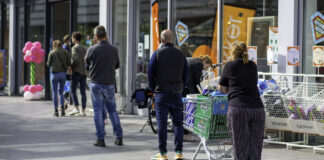 Viele Kunden stehen draußen vor einem Supermarkt oder Discounter Schlange. Einer schiebt bereits einen Einkaufskorb. Im Hintergrund kann man erkennen, dass es ein sonniger Tag ist.