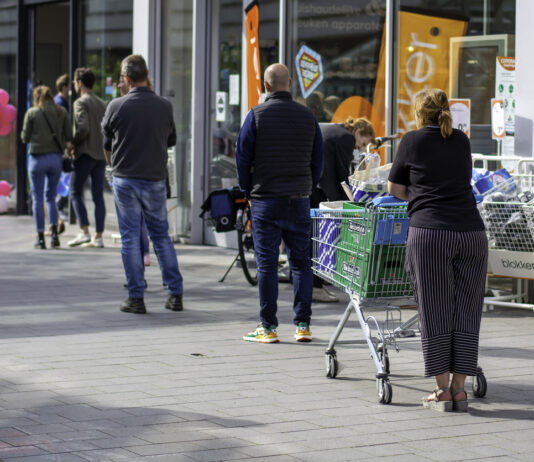 Viele Kunden stehen draußen vor einem Supermarkt oder Discounter Schlange. Einer schiebt bereits einen Einkaufskorb. Im Hintergrund kann man erkennen, dass es ein sonniger Tag ist.