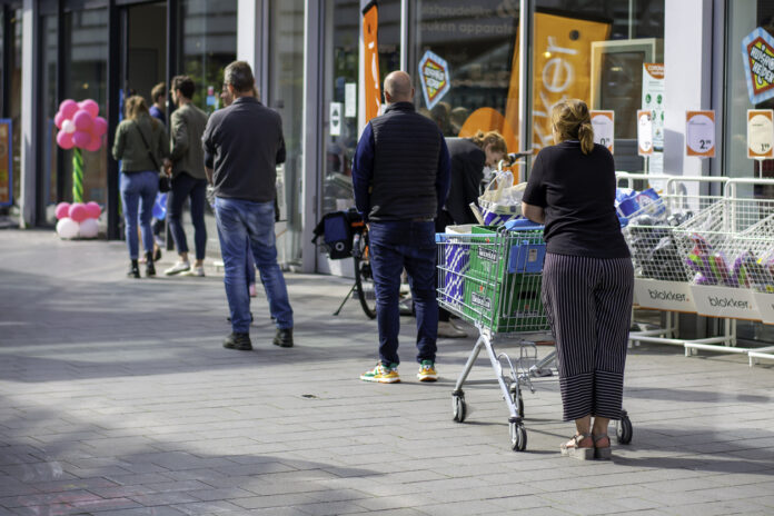 Viele Kunden stehen draußen vor einem Supermarkt oder Discounter Schlange. Einer schiebt bereits einen Einkaufskorb. Im Hintergrund kann man erkennen, dass es ein sonniger Tag ist.
