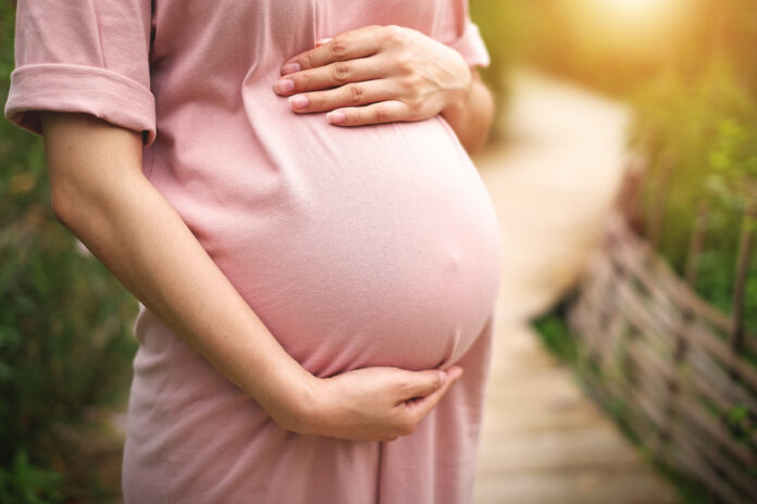 Eine Frau hält sich den dicken Babybauch. Sie steht im rosa Baumwollkleid auf einem hölzernen Pfad. Die Schwangerschaft steht auch für Lebensqualität und immer häufiger auch für eine überwundene Endometriose.