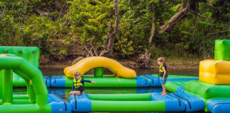 Zwei Kinder springen auf einer Hüpfburg, die sich auf dem Wasser befindet. Die Hüpfburg auf dem Badesee ist perfekt für einen Ausflug im Sommer.