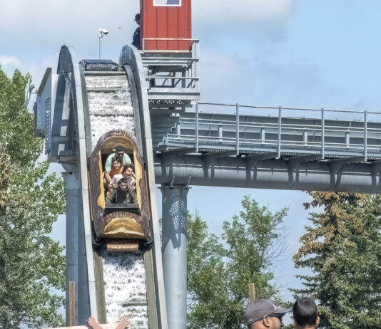 Menschen fahren im Freizeitpark auf einer Wasserbahn nach unten. Kinder sitzen in einem Wagen, der wie ein Boot aussieht. Im Hintergrund sind leicht bewölkter blauer Himmel und einige Bäume zu sehen.