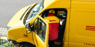 Ein Paketbote ist gerade dabei, sich wieder in sein Postauto zu setzen. Vorn auf dem Beifahrersitz sieht man einige Pakete und Päckchen liegen.