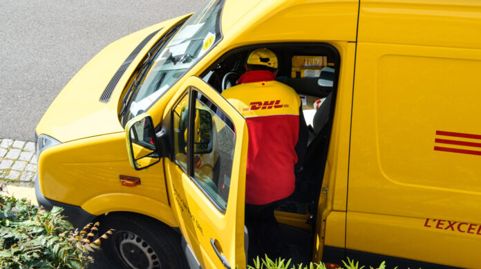 Ein Paketbote ist gerade dabei, sich wieder in sein Postauto zu setzen. Vorn auf dem Beifahrersitz sieht man einige Pakete und Päckchen liegen.