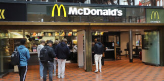 Eine lange Warteschlange vor einem McDonald's-Restaurant. Viele Menschen reihen sich an, teilweise noch vor dem Eingangsbereich, um an der Kasse der McDonald's-Filiale ihre Bestellung für Burger Pommes und Co. aufzugeben.