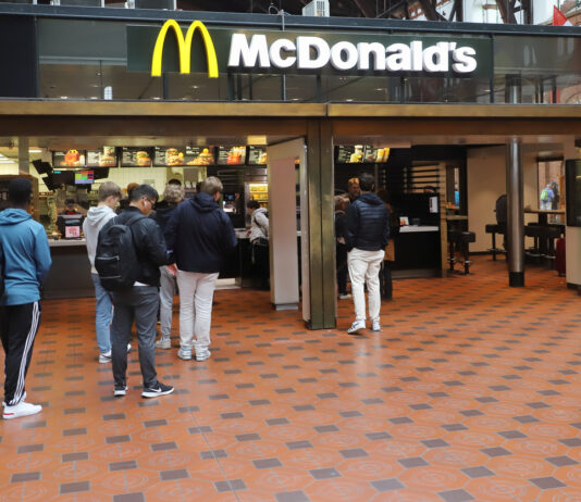 Eine lange Warteschlange vor einem McDonald's-Restaurant. Viele Menschen reihen sich an, teilweise noch vor dem Eingangsbereich, um an der Kasse der McDonald's-Filiale ihre Bestellung für Burger Pommes und Co. aufzugeben.