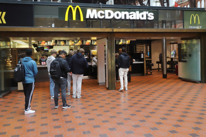 Eine lange Warteschlange vor einem McDonald's-Restaurant. Viele Menschen reihen sich an, teilweise noch vor dem Eingangsbereich, um an der Kasse der McDonald's-Filiale ihre Bestellung für Burger Pommes und Co. aufzugeben.