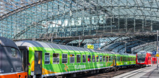 Ein grüner Flixtrain hält in einem Bahnhof. Daneben steht ein Zug der Deutschen Bahn. Die Sonne scheint durch das verglaste Dach des Bahnhofs.