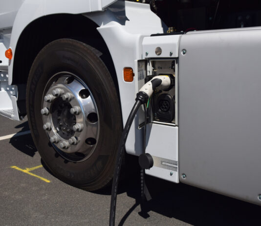 Die Großaufnahme eines Reifens und einer Ladebüchse von einem weißen E-Lkw. Ein schwarz-weißes Ladekabel steckt gerade in dem elektrischen Lastkraftwagen, das seinen Akku wieder neu voll auflädt.