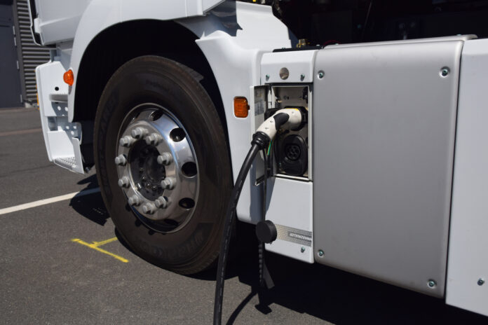 Die Großaufnahme eines Reifens und einer Ladebüchse von einem weißen E-Lkw. Ein schwarz-weißes Ladekabel steckt gerade in dem elektrischen Lastkraftwagen, das seinen Akku wieder neu voll auflädt.