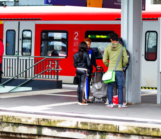 Fahrgäste am Bahngleis vor einem Zug der Deutschen Bahn. Eine Gruppe aus vier Menschen mit Rollkoffer wartet am Bahndamm. Links von ihnen steht ein Mann, alleine mit Handy und Fahrrad. Im Hintergrund steht ein roter Regionalexpress.