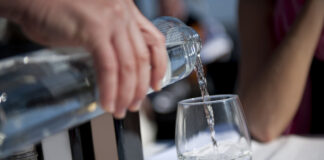 Mineralwasser oder Tafelwasser wird in ein auf weißer Tischdecke stehendes Glas eingegossen. Im Hintergrund sieht man den Arm eines Gastes aufgestützt auf dem Tisch.