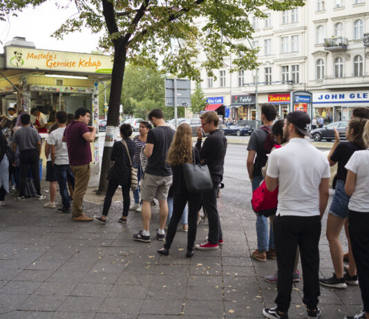 Menschenschlange vor einem türkischen Imbiss. Die Kunden warten darauf, ihren Döner bestellen zu können. Die Gäste stehen in einer schier endlosen Schlange auf dem Bordstein. Im Hintergrund prangt das Zeichen "Mustafas Gemüsedöner".