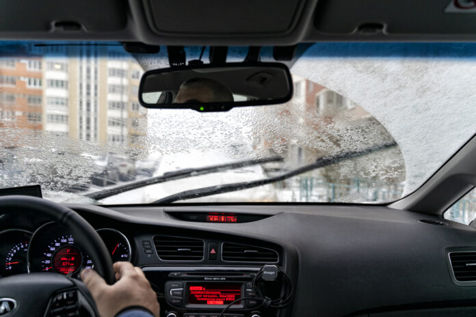 Die Innenansicht einer Autoscheibe bei tiefen Temperaturen im Herbst oder im Winter. Auf der Scheibe liegt ein wenig Frost oder Schnee. Ein Hand liegt am Lenkrad.