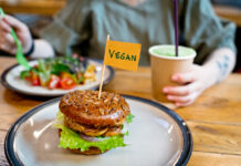 Eine Frau isst einen Salat und vor ihr auf einem weiteren Teller befindet sich ein Veggie Burger. Sie ernährt sich offensichtlich vegan und trinkt aus einem Coffee-to-go-Becher.