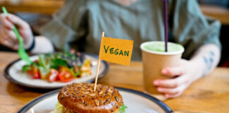 Eine Frau isst einen Salat und vor ihr auf einem weiteren Teller befindet sich ein Veggie Burger. Sie ernährt sich offensichtlich vegan und trinkt aus einem Coffee-to-go-Becher.